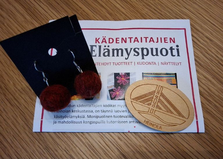 Puodin esite sekä Heinola-magneetti ja tummanpunaiset pyöreät lankakeräkorvakorut